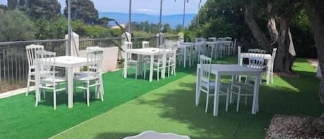 An outdoor seating area with white tables and chairs arranged on artificial grass, surrounded by trees and a clear sky.