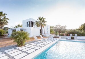 Une villa moderne aux murs blancs, dotée d’une piscine et entourée de palmiers et d’un jardin.