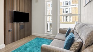 A living room with a flat-screen TV mounted on a wooden wall, a grey sofa with patterned cushions, a blue rug, and large windows offering a view of buildings outside. - Comfy Modern 2-Bed Notting Hill Portobello Apt (London)