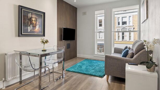 A modern living room with a glass table, a sofa, a blue rug, and a Buddha painting on the wall. - Comfy Modern 2-Bed Notting Hill Portobello Apt (London)