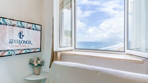 Huone, josta avautuu näkymä merelle, seinällä oleva kehystetty kyltti, jossa lukee ”SPERLONGA RESORT”, ja pieni valkoinen hylly, jonka päällä on maljakko.