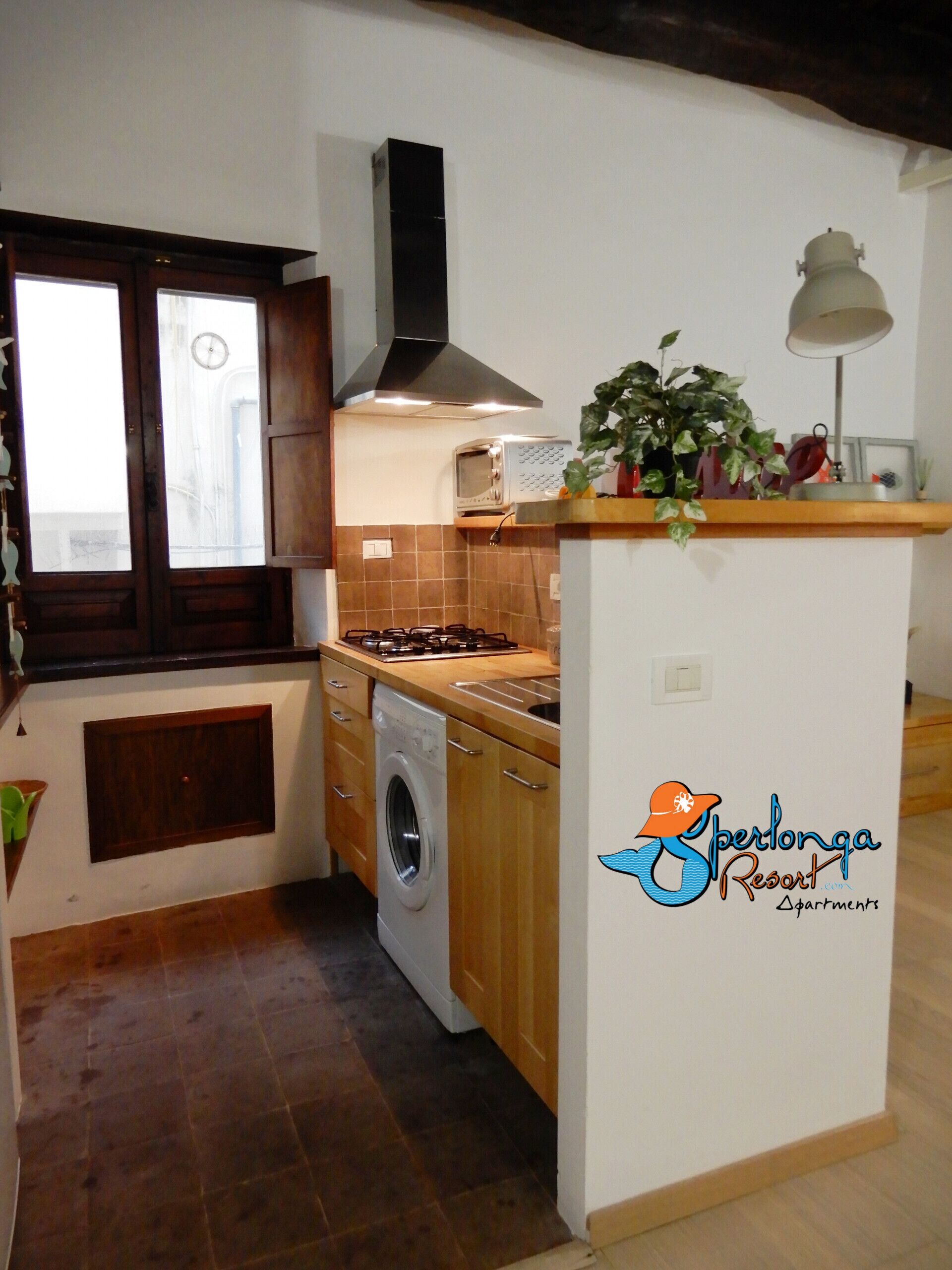 A compact kitchen with a washing machine, a stove, and a microwave. There's a window with curtains, a plant on the counter, and a logo for 'Sperlonga Resort'.