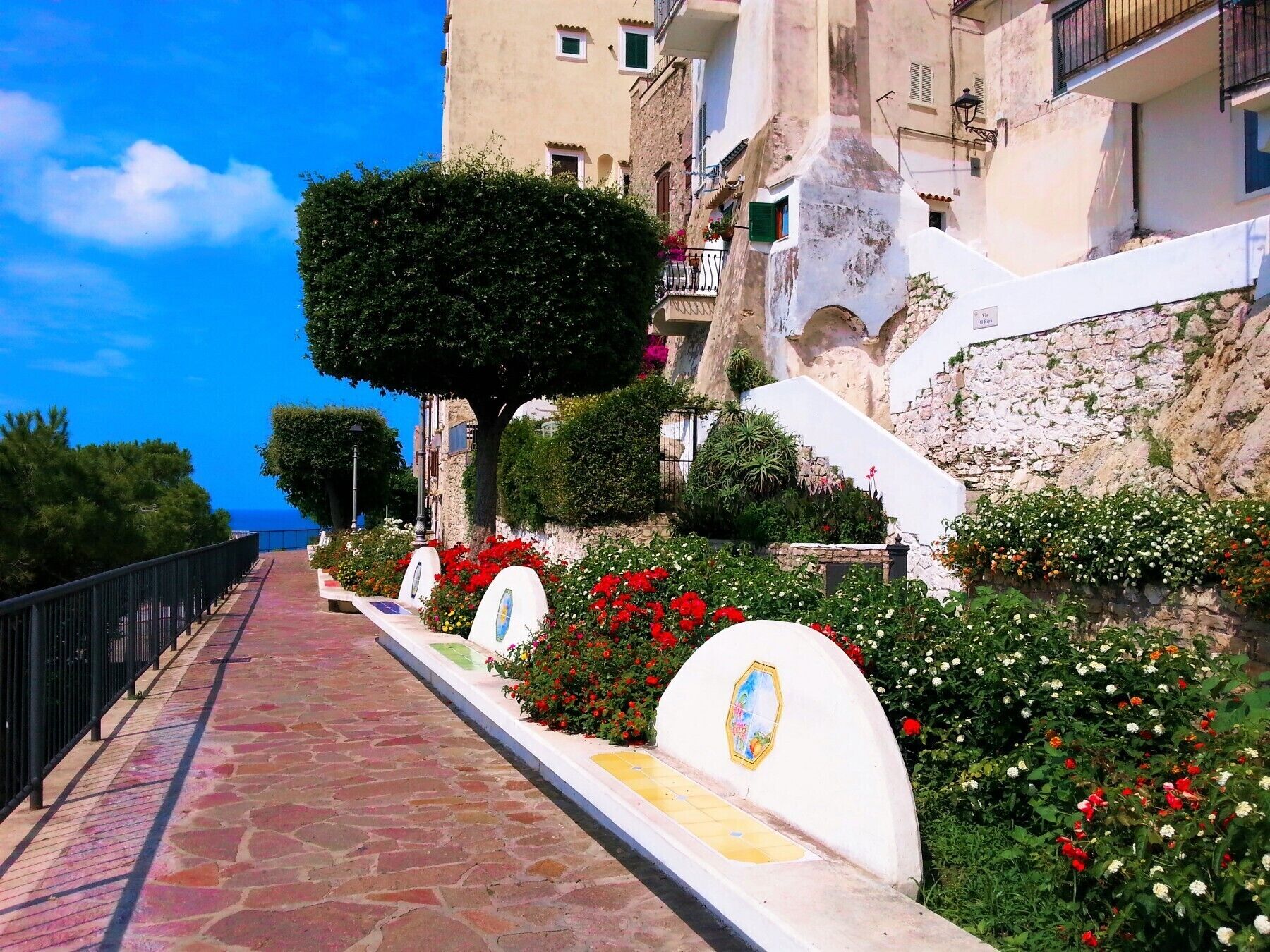 Un sentier pavé de pierres, bordé de fleurs aux couleurs vives et d’un arbre, mène à un bâtiment doté de balcons et d’un mur en pierre.