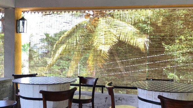A covered outdoor seating area with wooden chairs and tables, a view of palm trees through a woven screen, and a tiled floor. - Blue Pillars Kandy (Kandy)