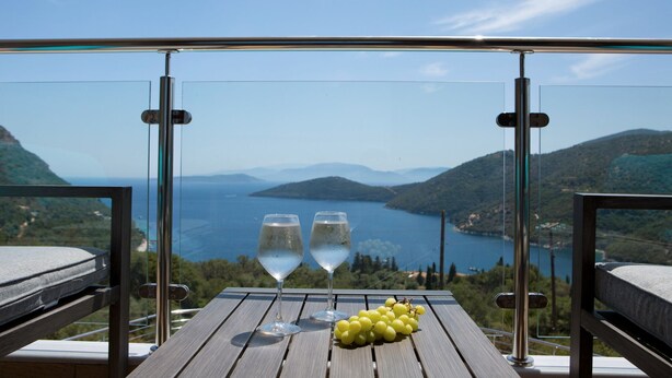 Un balcón con dos vasos de agua y un racimo de uvas, con vistas a un paisaje pintoresco de un lago y montañas.