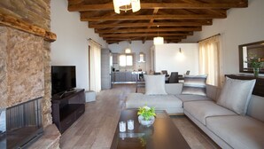 Una sala de estar con chimenea de piedra, vigas de madera en el techo, un sofá, una mesa de centro con un florero y una cocina al fondo.