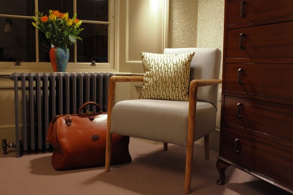 Een kamer met een radiator, een stoel met een gekleurd kussen, een bruine leren tas en een houten dressoir.