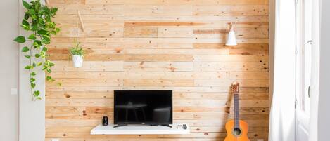 Una habitación con pared de madera, un televisor sobre un soporte blanco, una guitarra y una planta colgante.