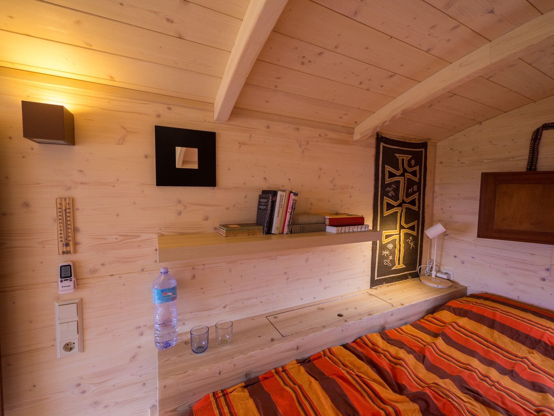 A cozy wooden cabin room with a bed, a shelf with books, a wall-mounted light, and a door with a decorative pattern.