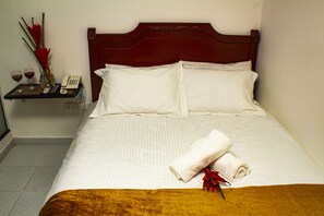 Un lit bien fait, avec du linge de lit blanc, un plaid marron et une serviette enroulée. Un vase avec des fleurs rouges et deux verres de vin rouge sont posés sur une petite table à côté du lit.