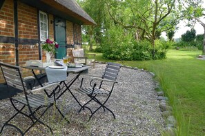 Ein rustikales Häuschen mit Strohdach, Backsteinfassade und Kiesterrasse. Zwei Metallstühle und ein Tisch mit kariertem Tischläufer stehen bereit.