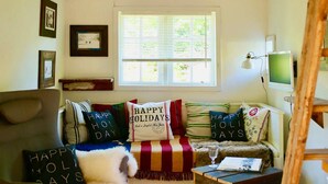 A cozy room with a sloped ceiling, a window with blinds, a sofa with holiday-themed pillows, a chair, a small wooden table with a glass, and a TV on a stand. - Kl. Reetkate f. 2 pers. / WLAN / sauna / garden / a few minutes to the beach (Behrensdorf)