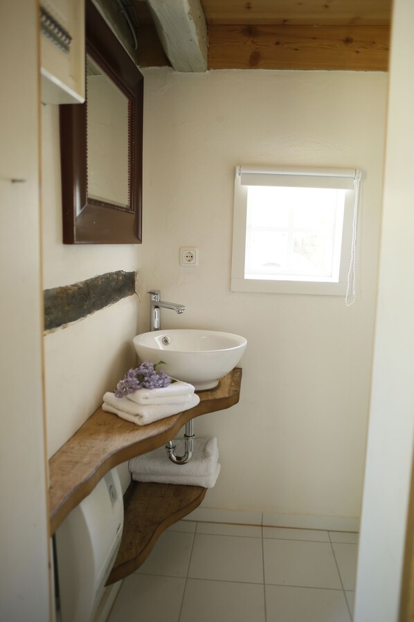 Un baño con lavamanos blanco, tocador de madera, espejo y ventana con persianas.