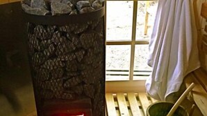 A wood-burning stove with a fire, a window with a view of greenery, and a wooden bench with a towel hanging on it. - Kl. Reetkate f. 2 pers. / WLAN / sauna / garden / a few minutes to the beach (Behrensdorf)