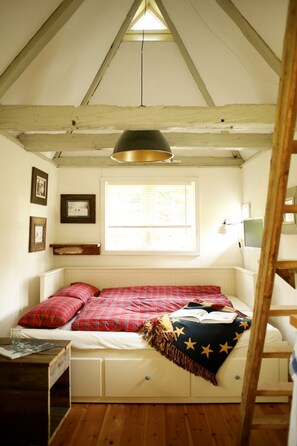 Ein gemütliches Schlafzimmer im Dachgeschoss mit schrägen Wänden, einem Fenster mit Jalousien, einem Bett mit roter Karo-Bettwäsche, einer Stern-Decke und einer Holzleiter.
