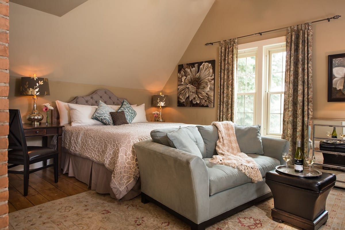 A bedroom with a bed, a sofa, a desk, and a chair. There are curtains on the windows, a painting on the wall, and a vase with flowers on the desk.