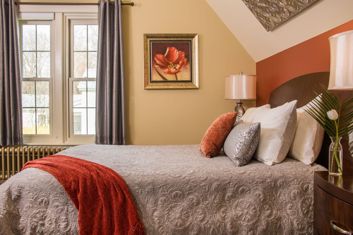 A neatly made bed with a patterned bedspread, a red throw blanket, and several pillows. There is a framed picture on the wall, a bedside lamp, and a vase with a plant on the nightstand.
