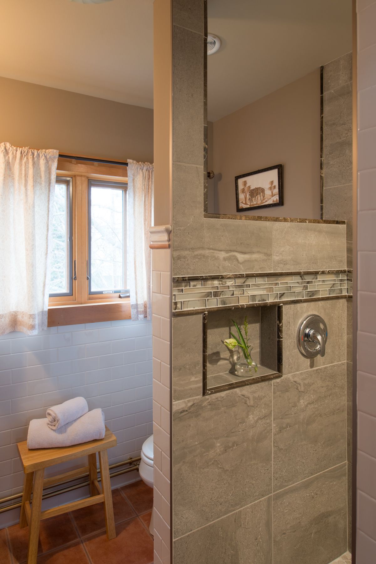 A bathroom with a shower area, a window with curtains, a towel on a stool, and a framed picture on the wall.