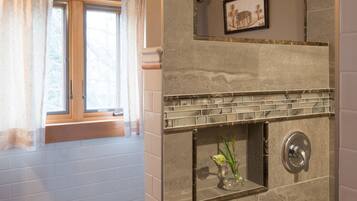 Une salle de bain avec un espace douche, une fenêtre avec des rideaux, une serviette posée sur un tabouret et un tableau encadré au mur.