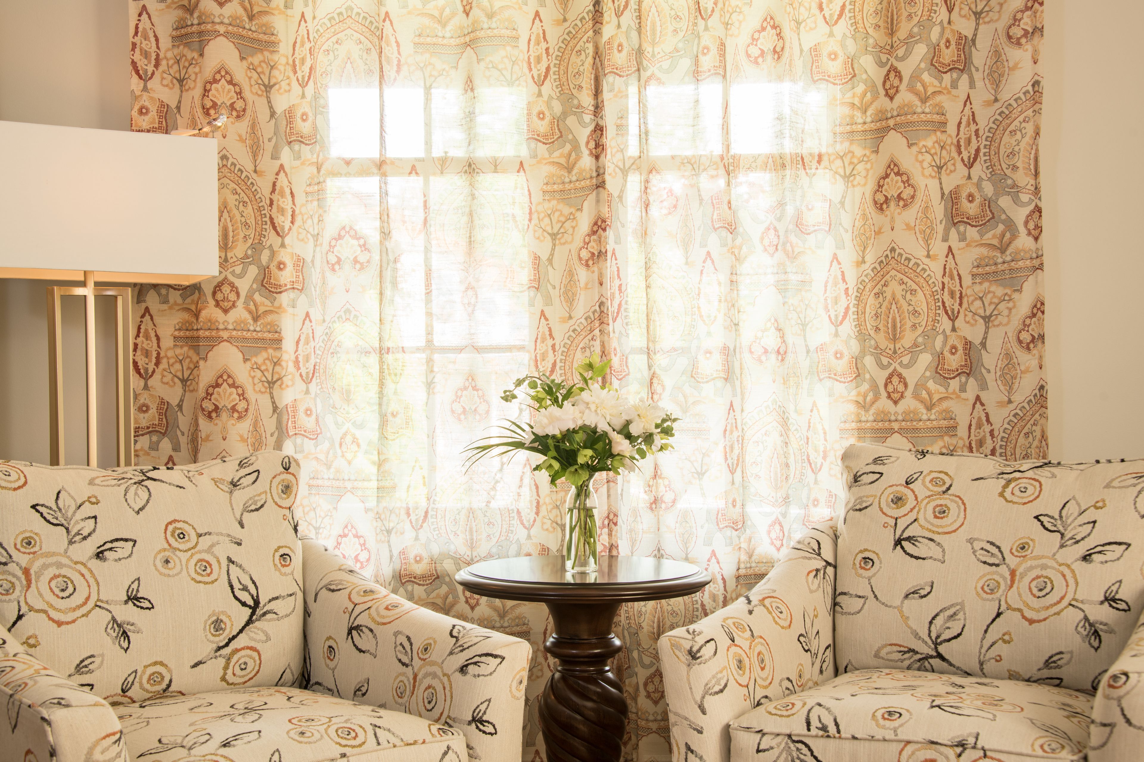 A room with two patterned armchairs, a small round table with a vase of flowers, and patterned curtains.