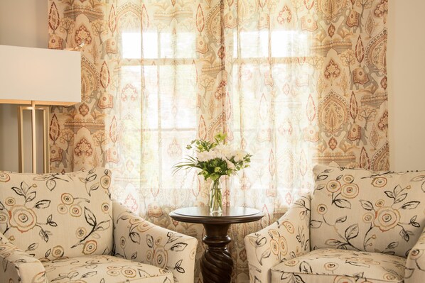 Ein Zimmer mit zwei gemusterten Sesseln, einem kleinen runden Tisch mit Blumenvase und gemusterten Vorhängen.