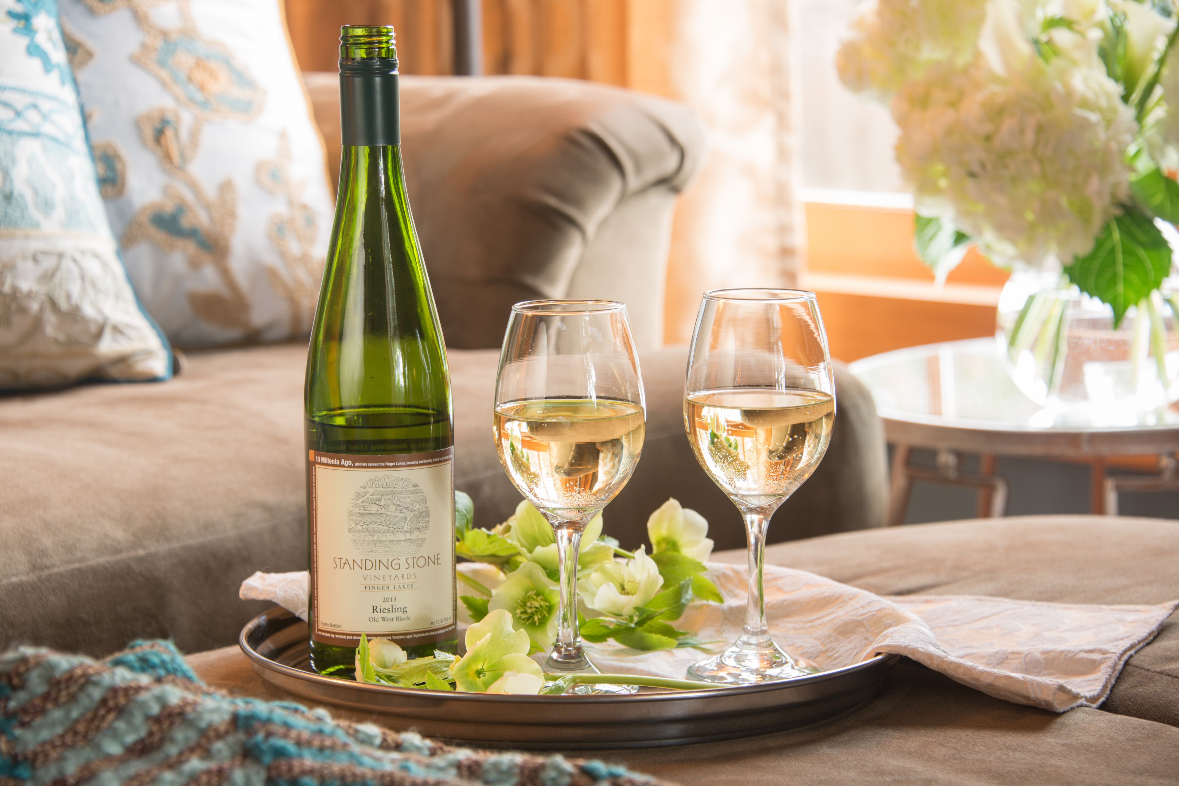 A bottle of wine, two glasses, and a tray with flowers on a table in a cozy room.