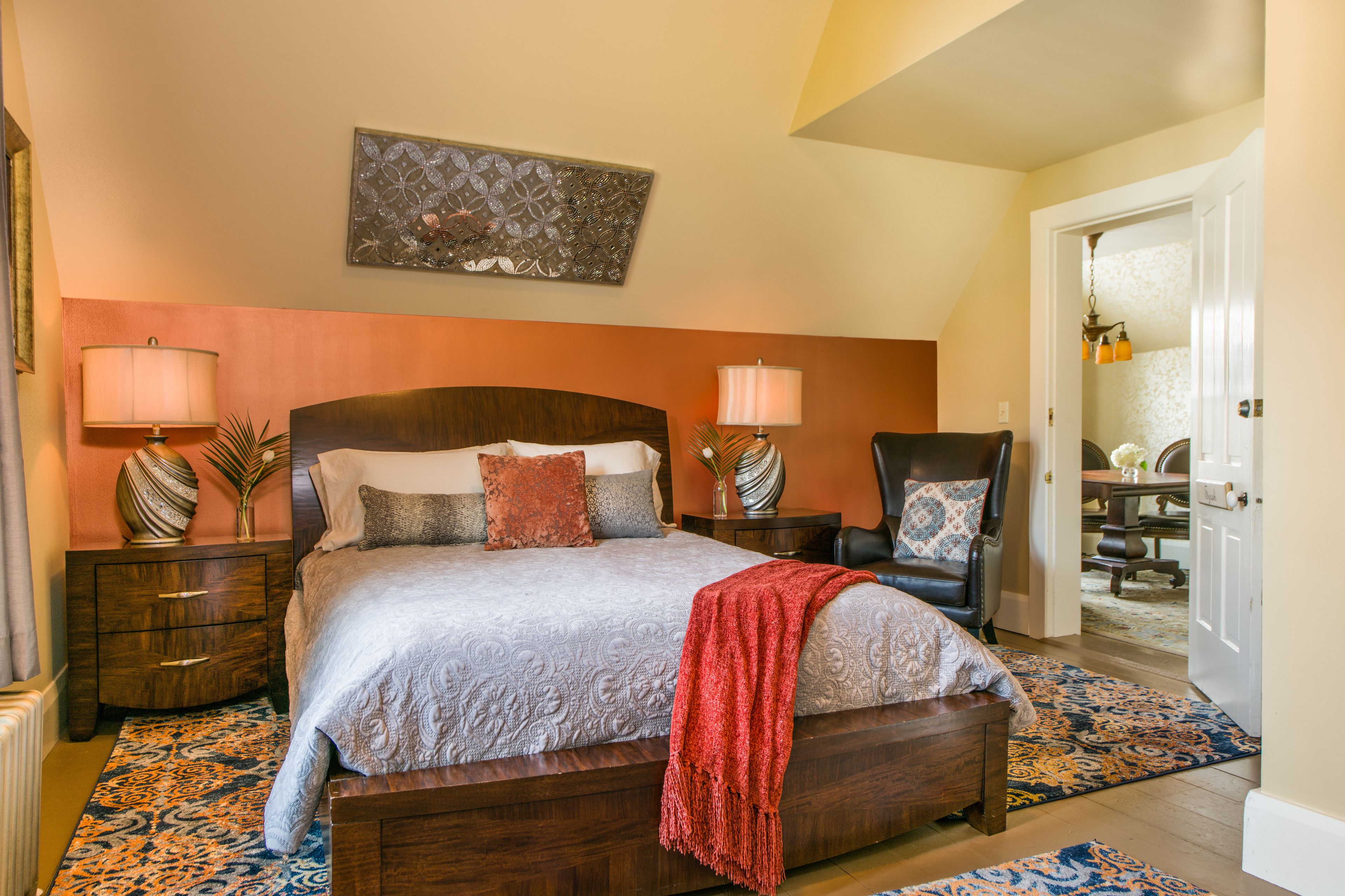 A bedroom with a large bed, two nightstands, a chair, and a door leading to a dining area.