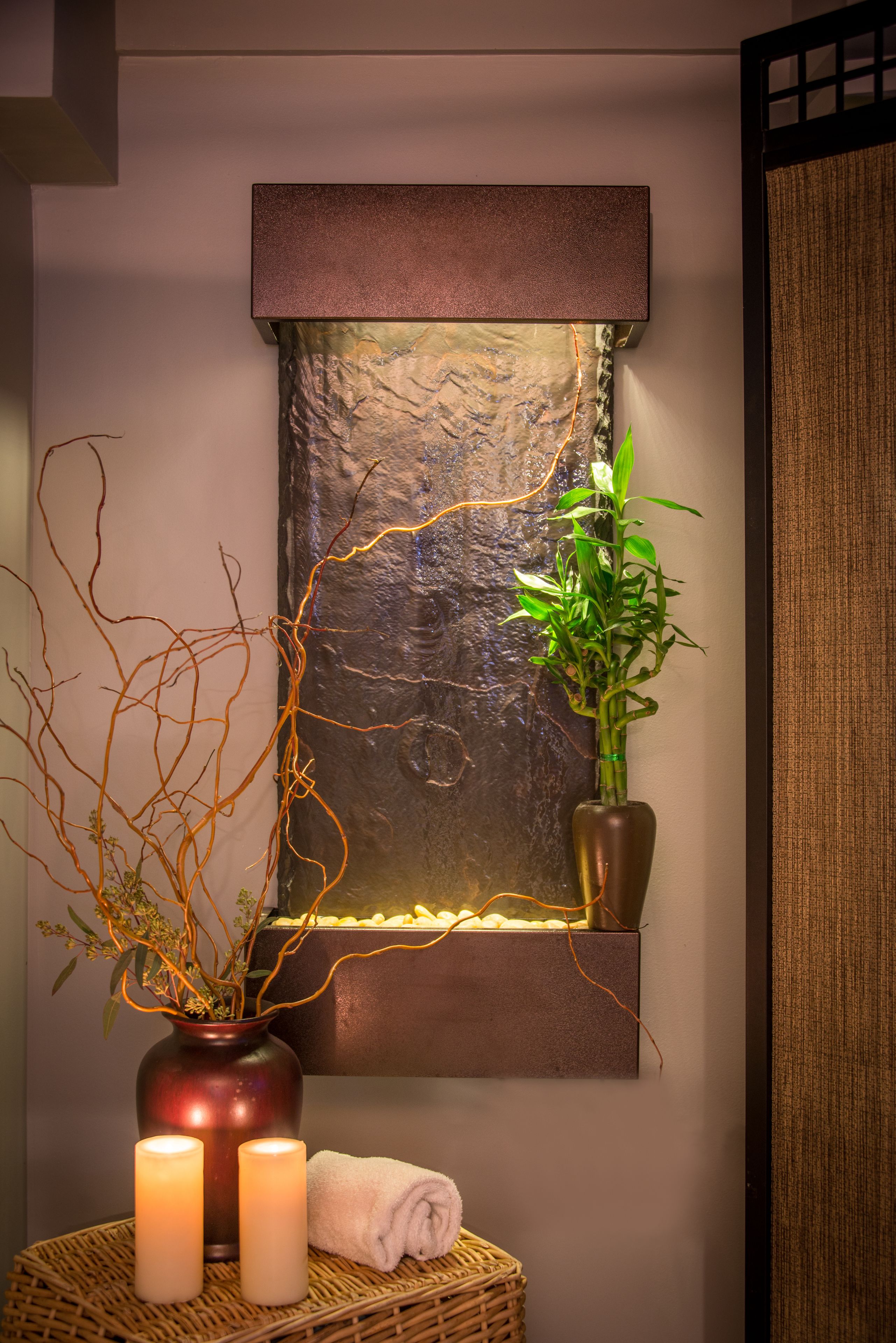 A wall-mounted water feature with a textured stone facade, a potted plant, and two candles on a woven surface.