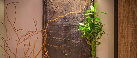 A wall-mounted water feature with a textured stone facade, a potted plant, and two candles on a woven surface.