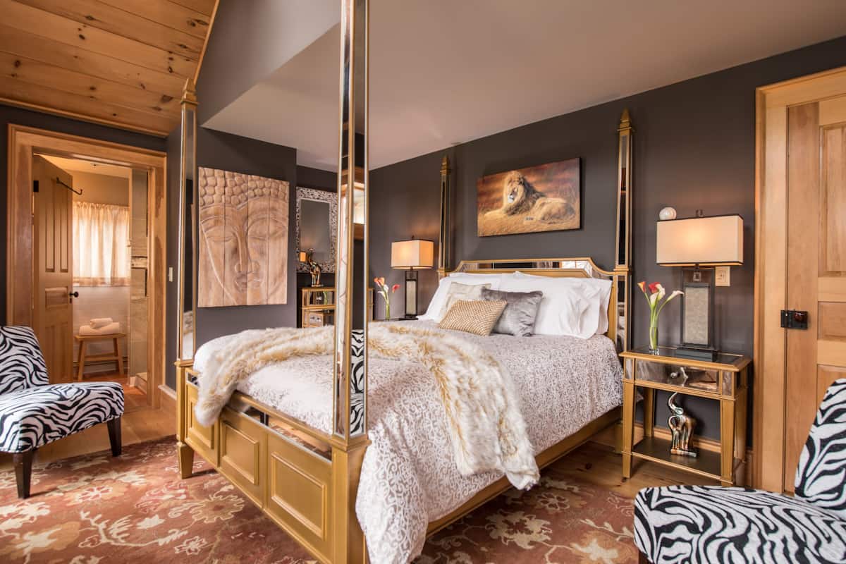 A four-poster bed with a white and gold patterned bedspread, a zebra-patterned armchair, a wooden nightstand with a vase of flowers, and a painting of a lion on the wall.