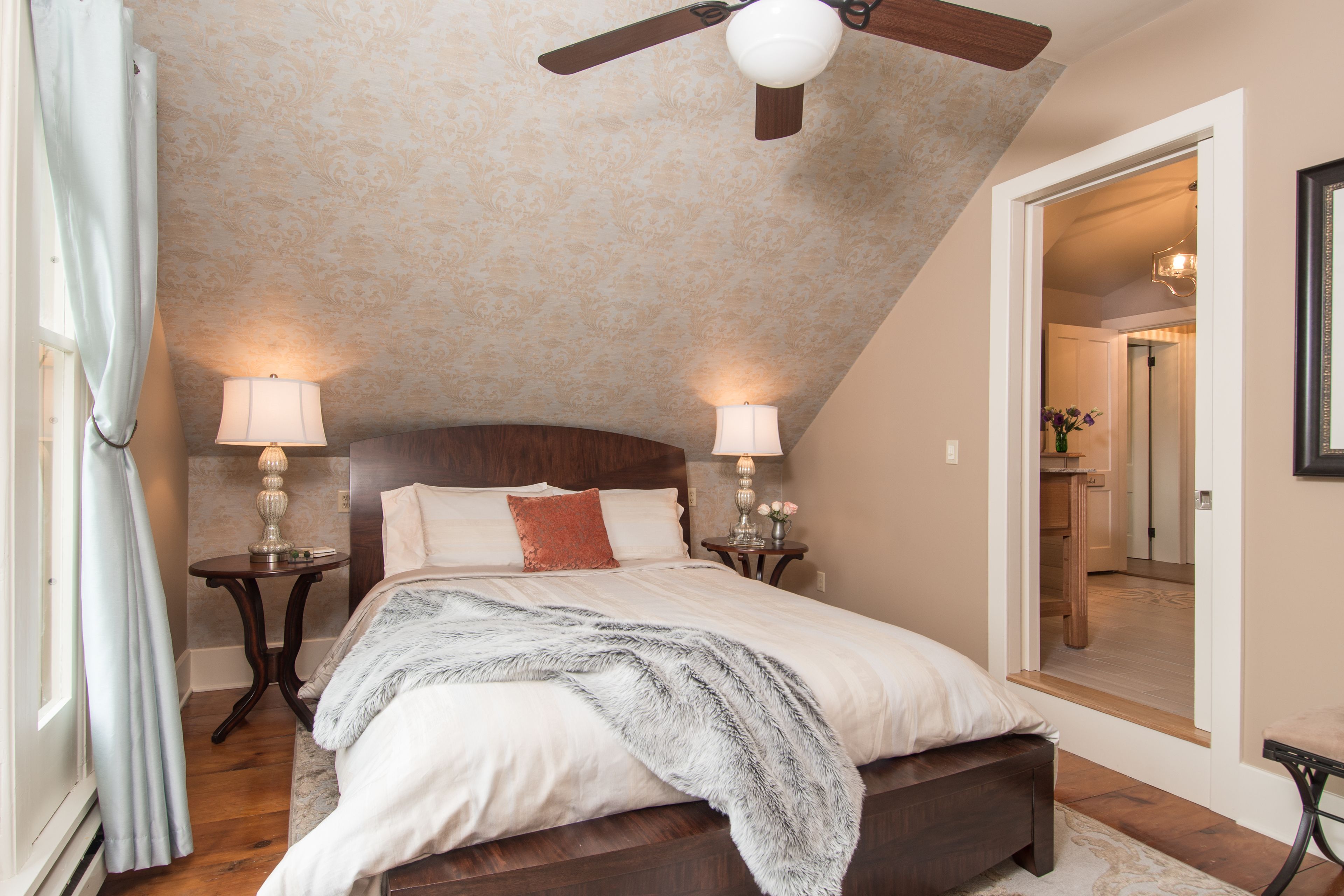 A bedroom with a sloped ceiling, a bed with a headboard, two bedside tables with lamps, and a mirror on the wall.