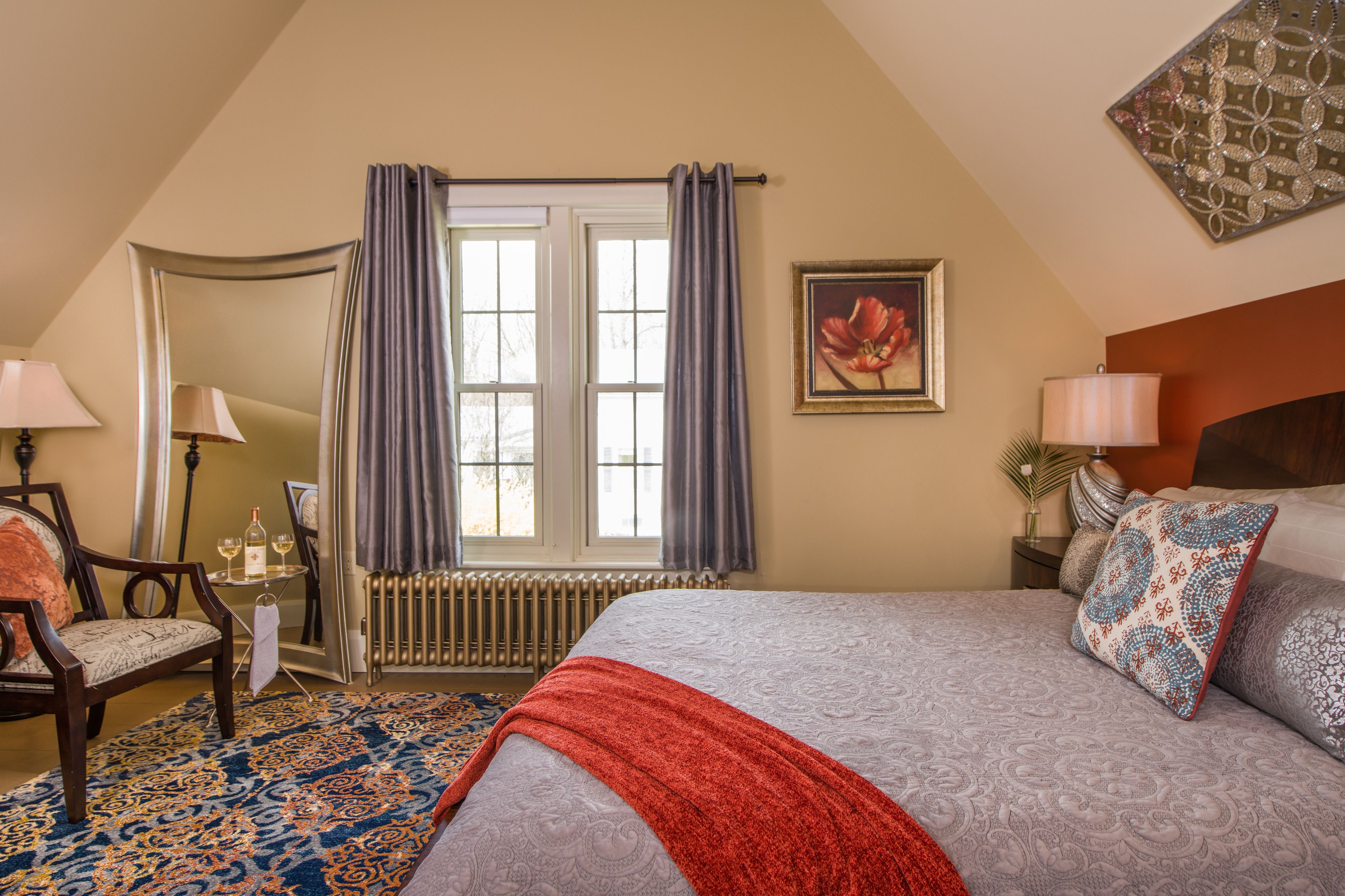 A bedroom with a large bed, a chair, a small table with a lamp, a radiator, and a window with curtains.