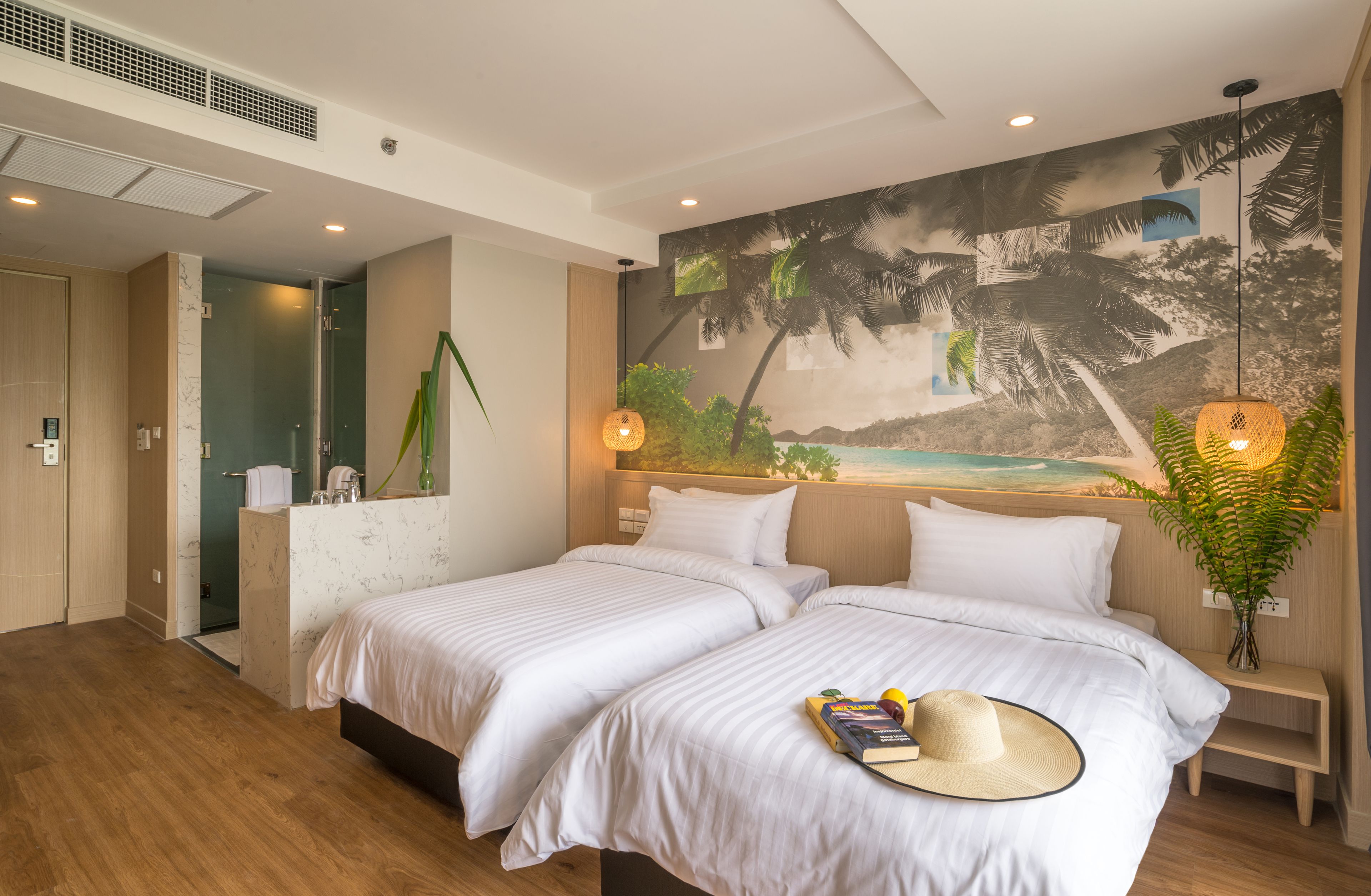 A hotel room with two beds, a mural of palm trees, a small plant, and a bedside table with a lamp.