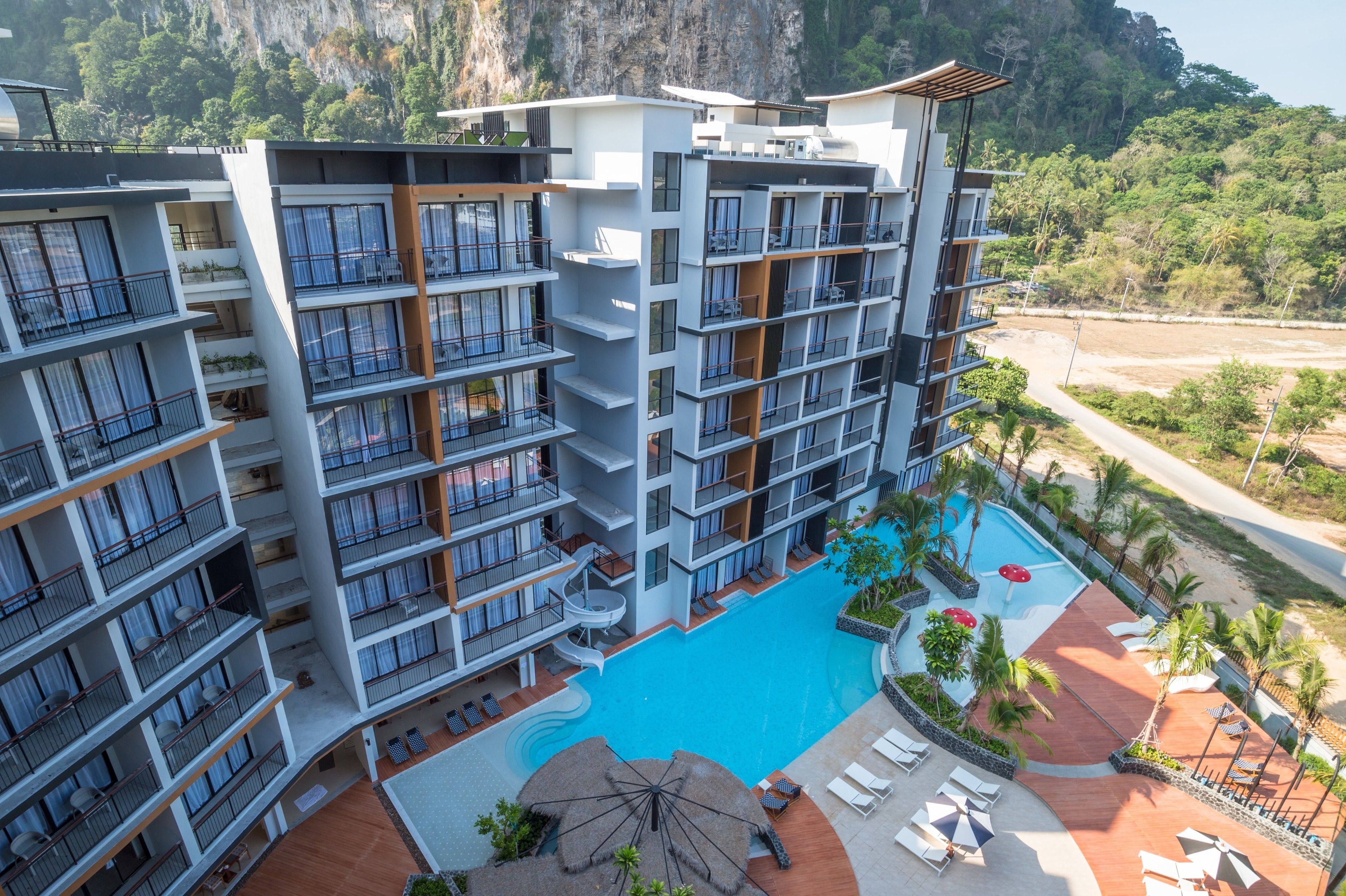 A multi-story hotel with balconies, a swimming pool, and a view of a mountainous landscape.