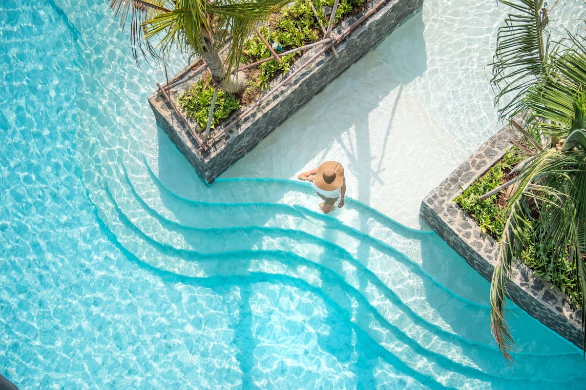 Une vue aérienne d’une piscine d’un bleu éclatant, où l’on aperçoit une personne debout près du bord, entourée de bordures en pierre et de verdure.