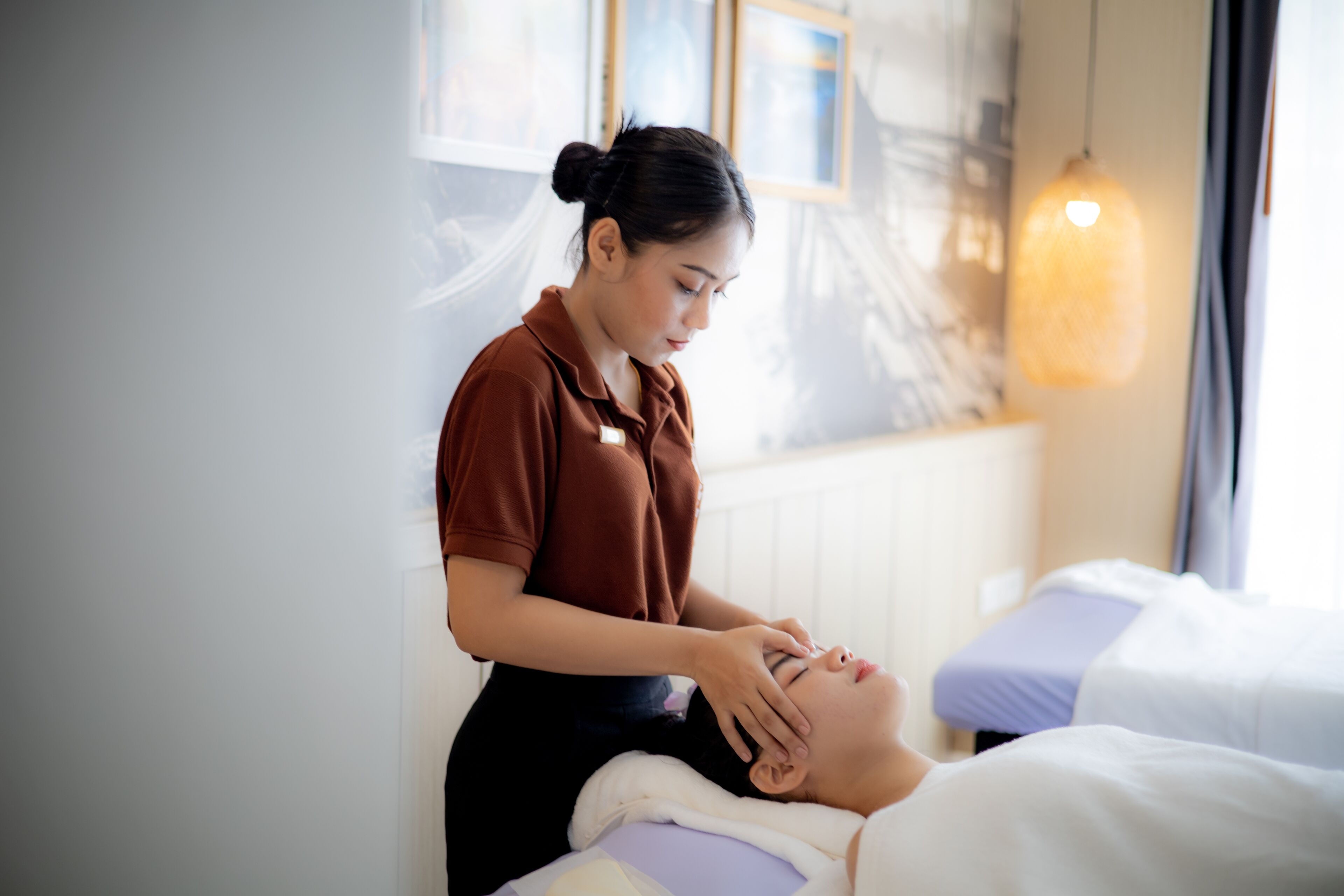 A person receiving a facial treatment in a room with a bed, a window with curtains, and a hanging light fixture.