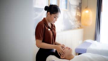 Una persona recibe un tratamiento facial en una habitación con una cama, una ventana con cortinas y un lámpara colgante.