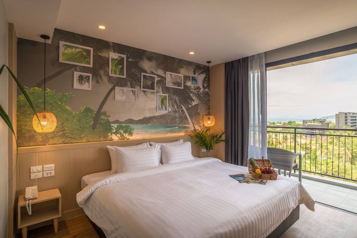 Une chambre d’hôtel avec un grand lit, une vue sur la ville, un balcon et une fresque murale aux motifs tropicaux.