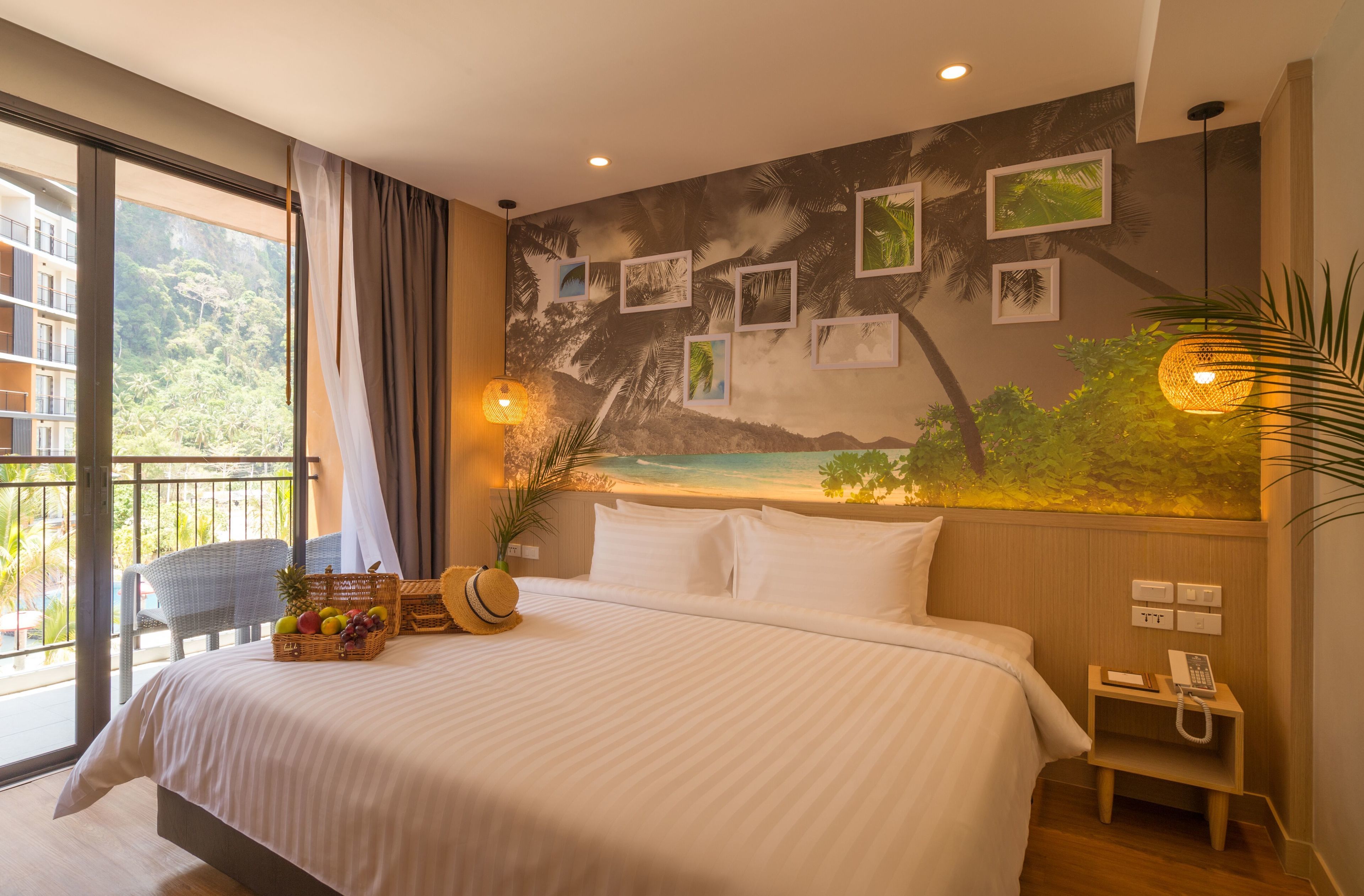 Habitación de hotel con una cama grande, un balcón con vistas a una montaña y árboles, una pared con cuadros enmarcados, y una mesita de noche pequeña con un teléfono.