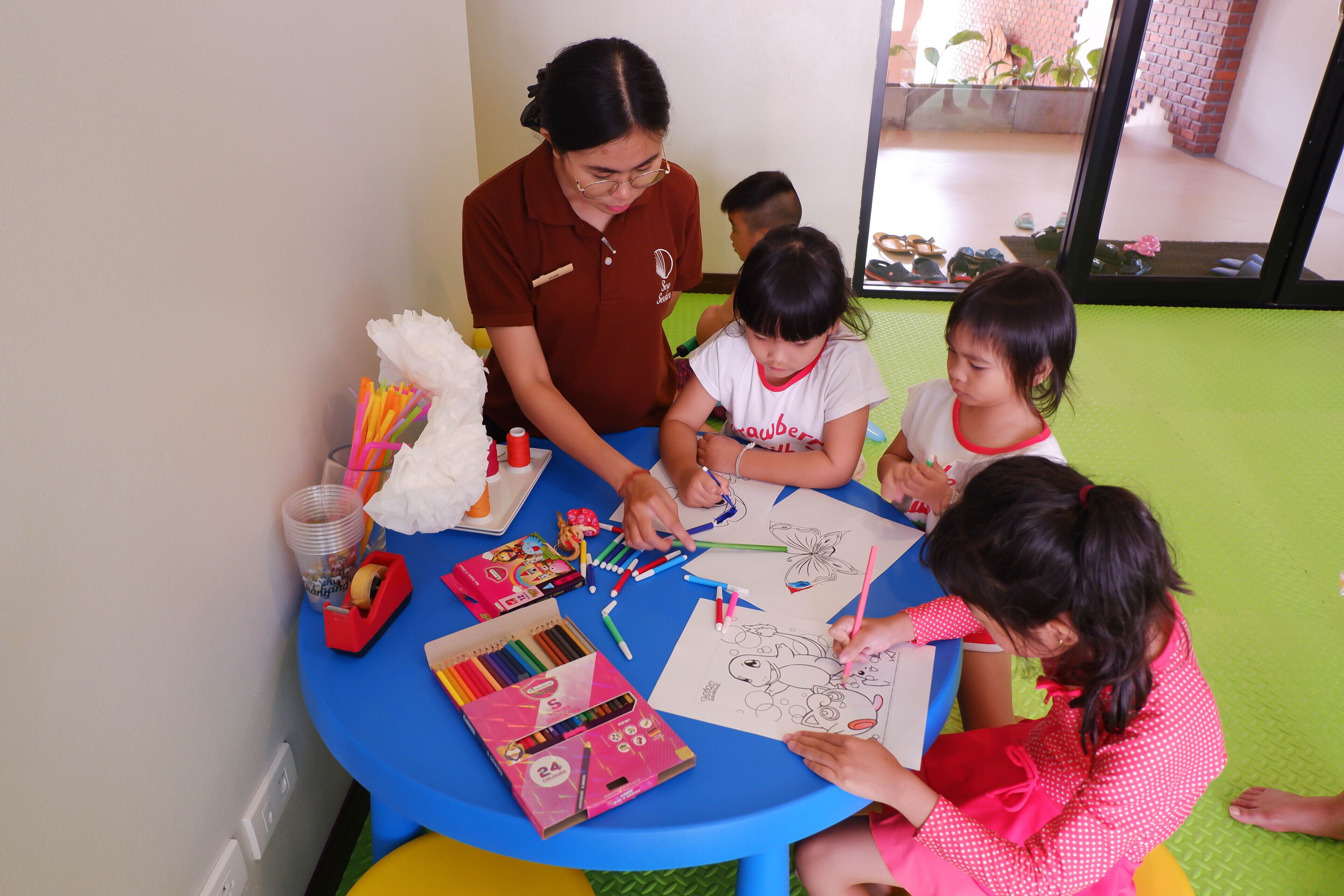 A group of children and an adult engaged in a drawing activity at a round table with art supplies.
