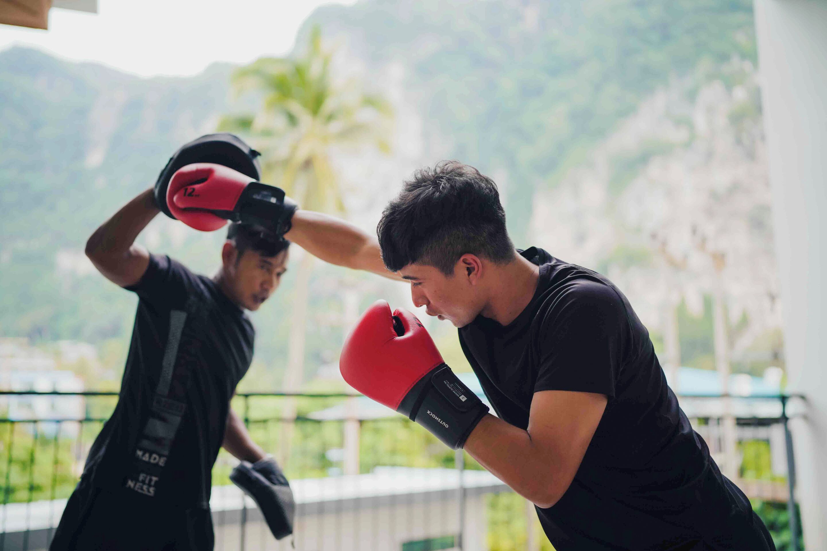 Two people in black athletic wear practicing boxing with red gloves, outdoors with a view of greenery and mountains in the background.