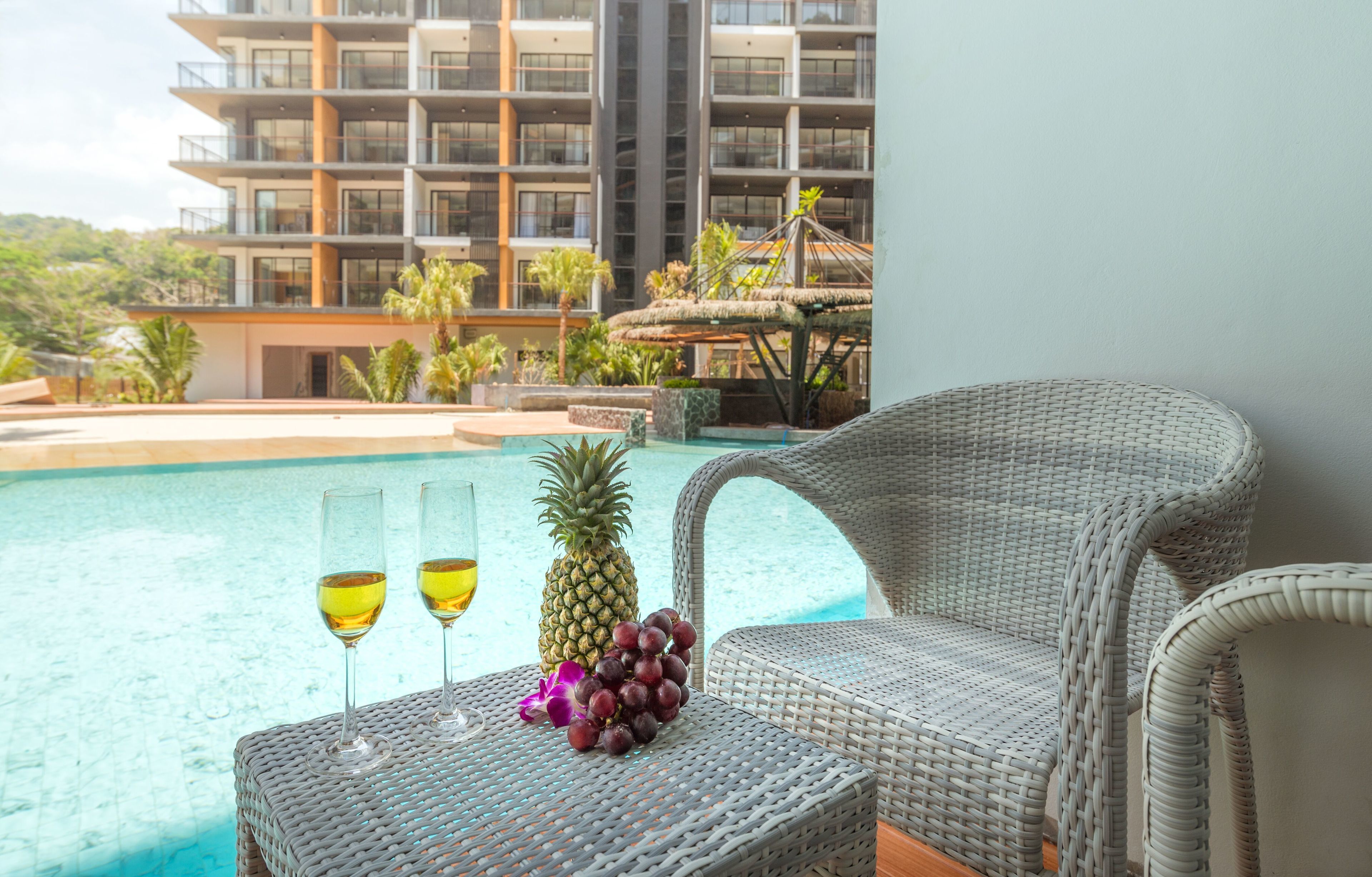 A wicker lounge chair by a pool with two glasses of white wine and a pineapple.