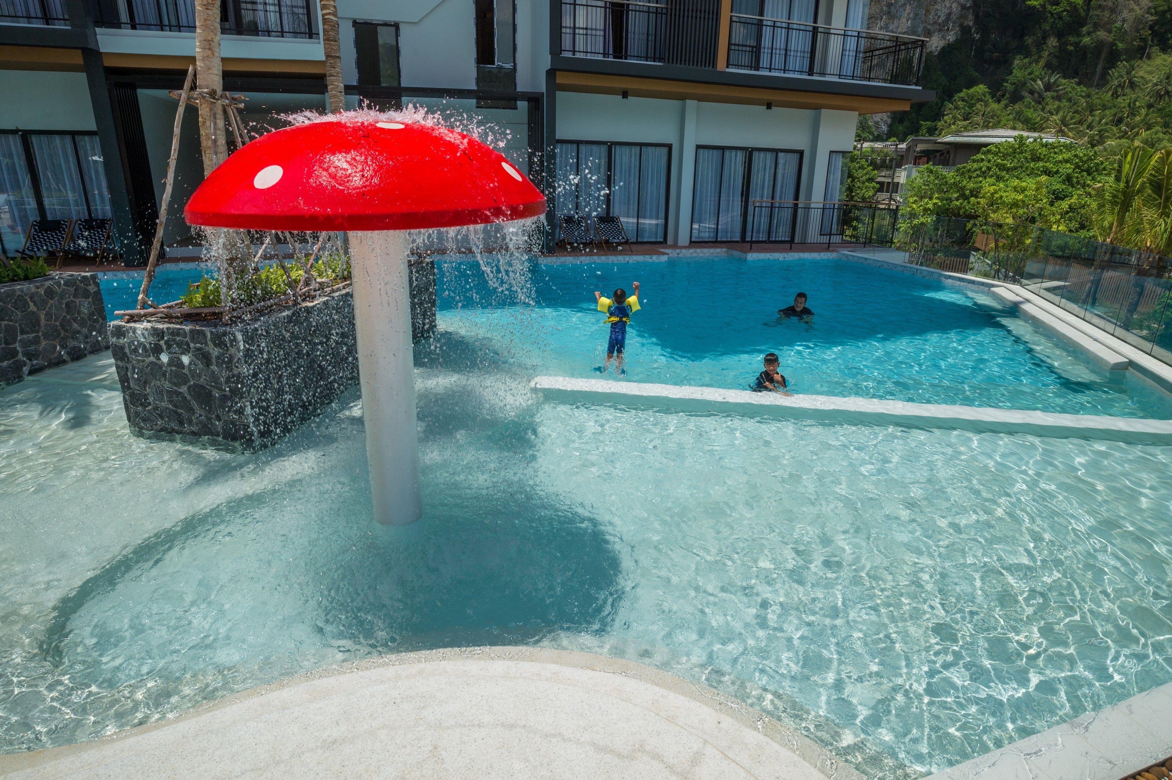 A swimming pool with a mushroom water feature, surrounded by stone planters and a building with balconies.