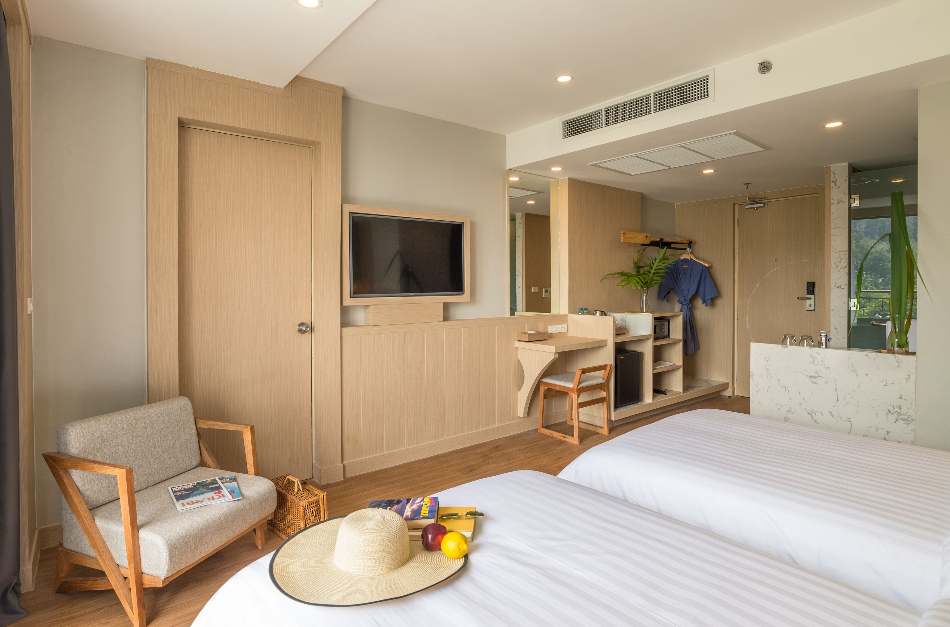 A modern hotel room with a large bed, a seating area with a magazine, a hat, and fruits, a desk with a computer, and a shower area.