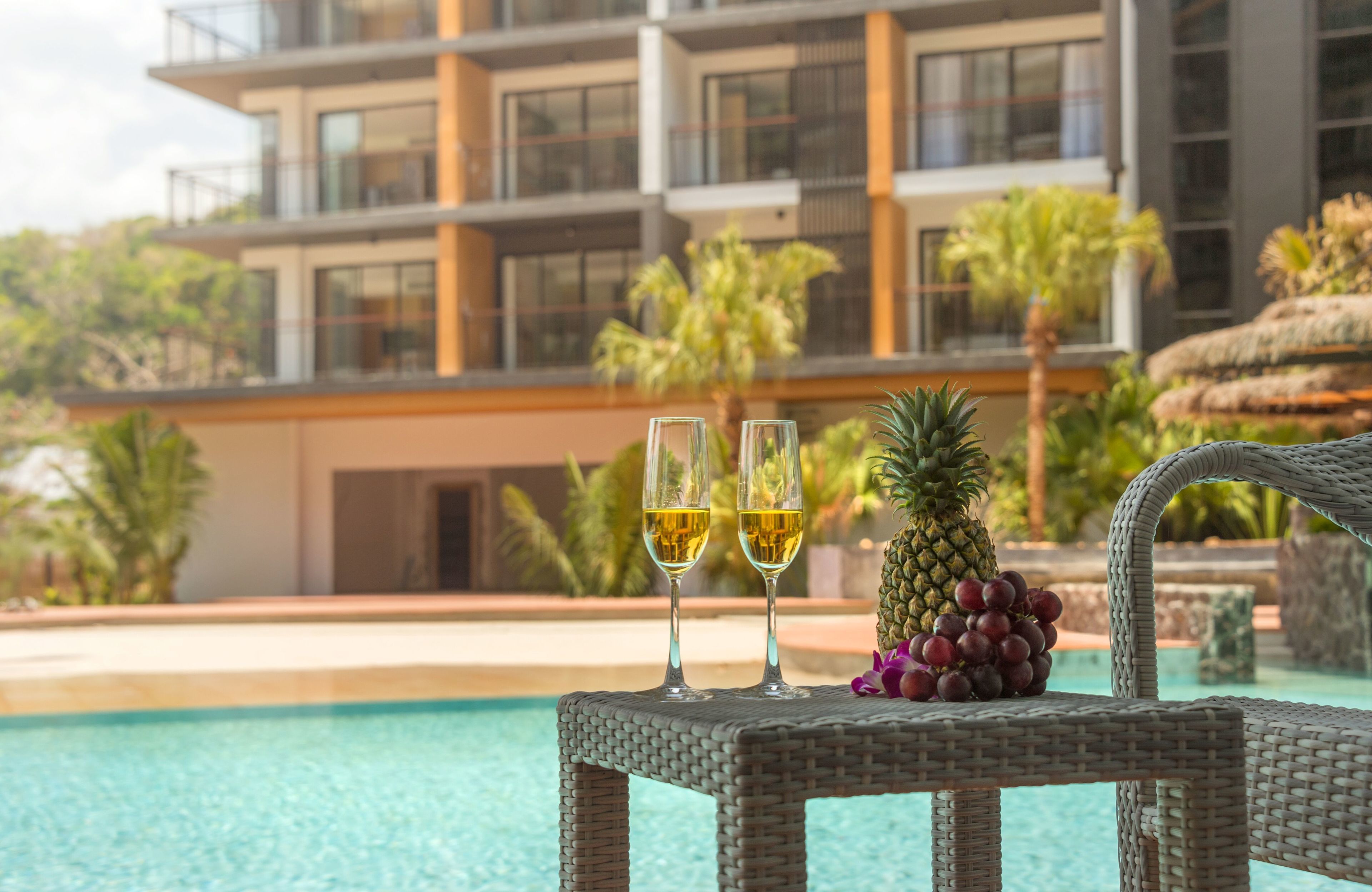 A poolside setting with two glasses of white wine, a pineapple, and grapes on a wicker table.