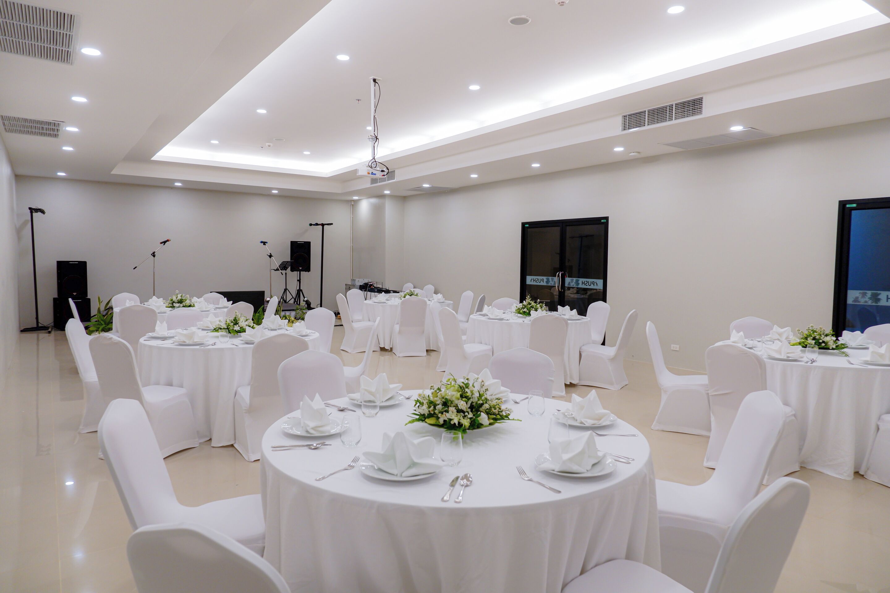 Un amplio y bien iluminado salón de banquetes con varias mesas redondas dispuestas para cenar, cada una con mantel blanco y centros de mesa florales.