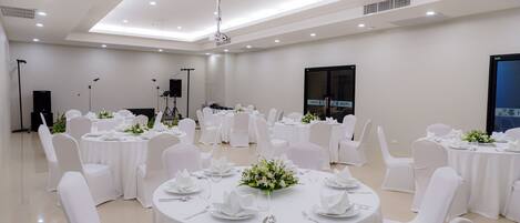 Un amplio y bien iluminado salón de banquetes con varias mesas redondas dispuestas para cenar, cada una con mantel blanco y centros de mesa florales.