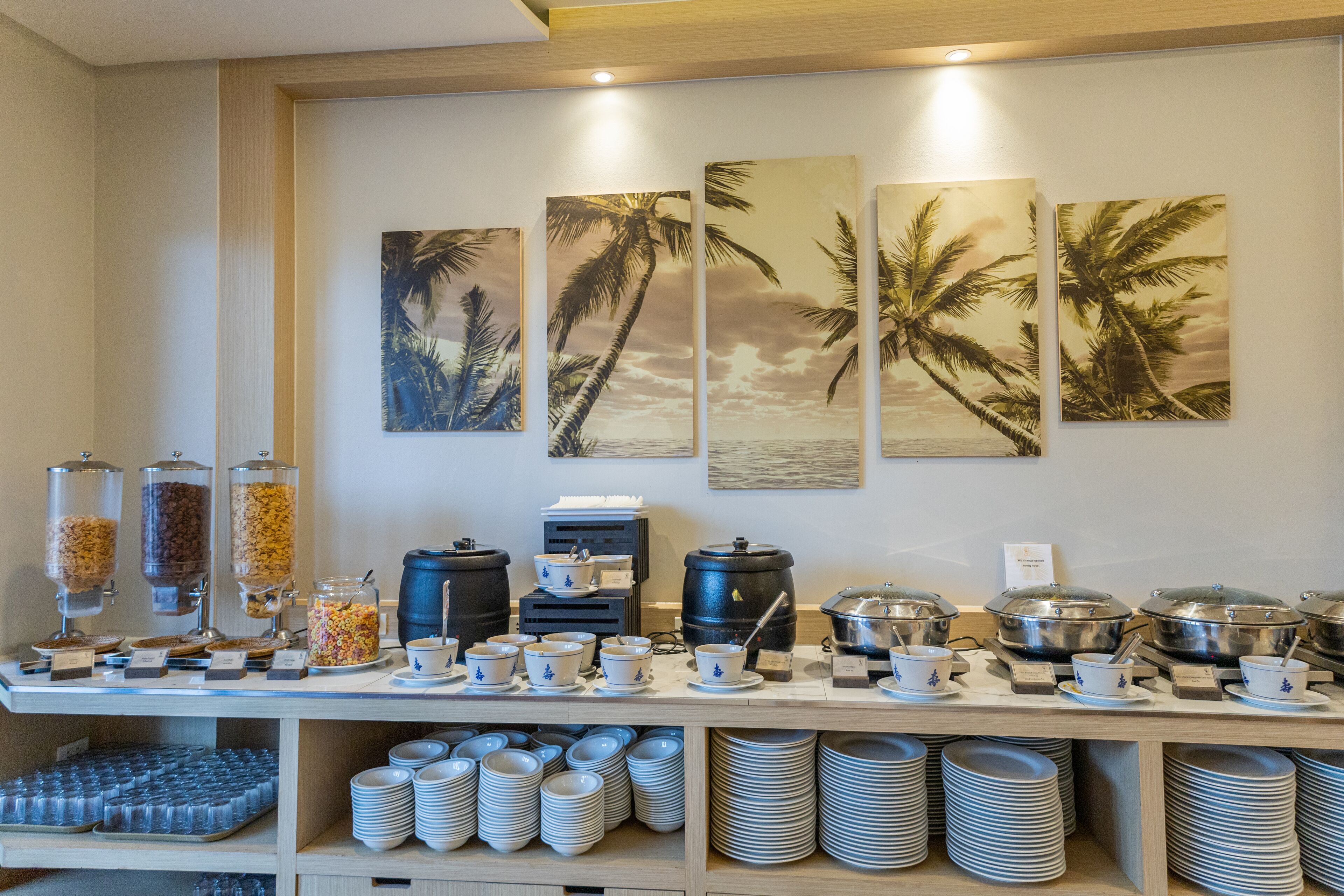 A buffet setup with various food containers, serving utensils, and a wall decorated with palm tree paintings.