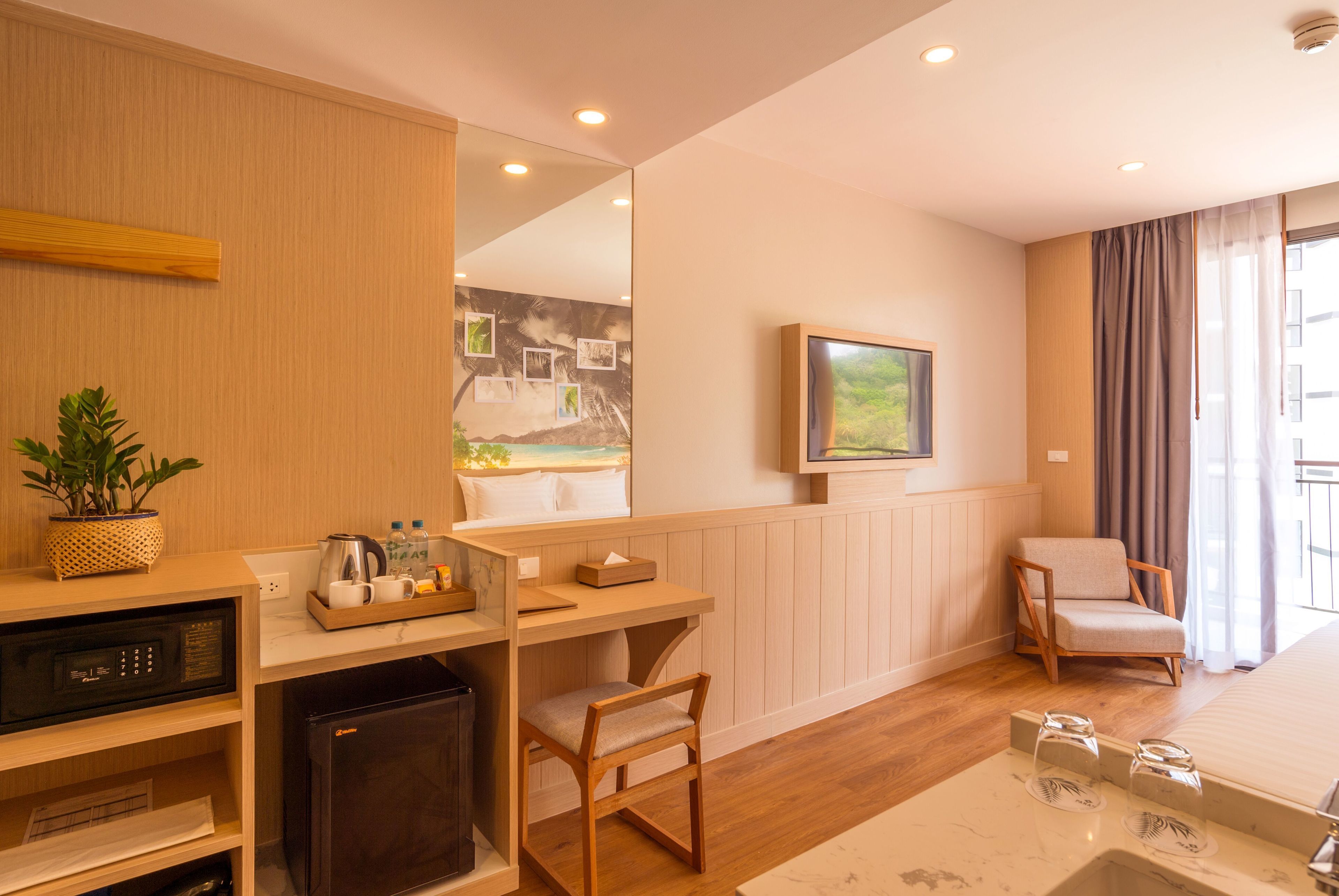 A modern hotel room with a wooden desk, a small refrigerator, a comfortable chair, a bed, and a large mirror.