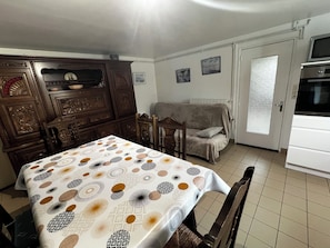 Un coin repas avec une table recouverte d’une nappe à motifs, un buffet en bois orné de sculptures décoratives, un canapé et un coin cuisine équipé d’un four à micro-ondes.